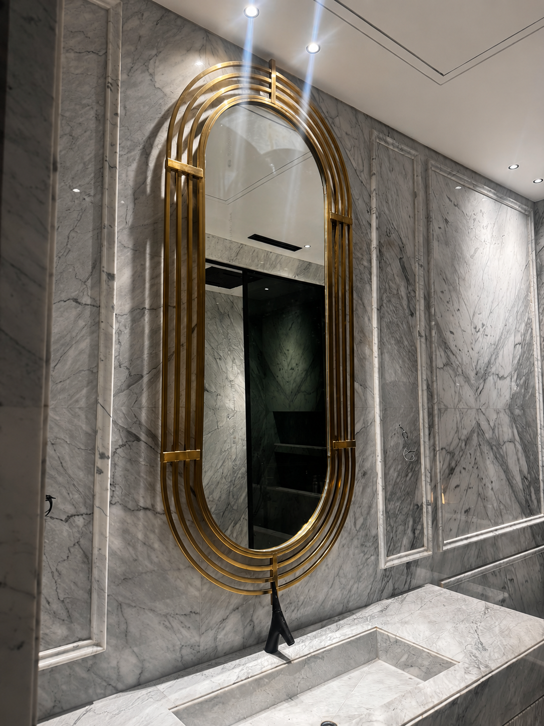 A bespoke elongated mirror framed with multiple curved gold PVD profiles, designed as a striking focal point against the grey marble wall.